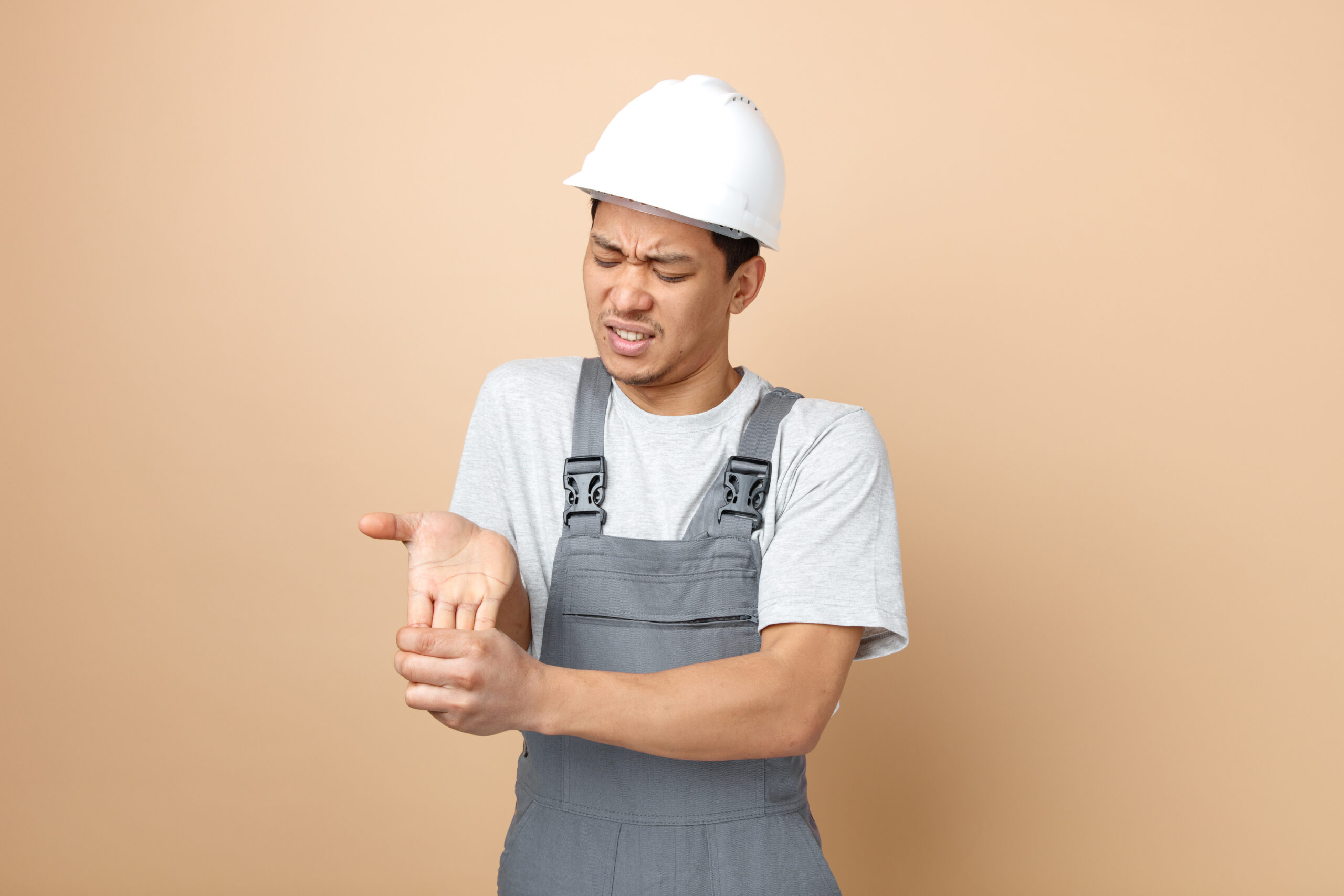 aching young construction worker wearing safety helmet and uniform holding hand with closed eyes isolated on creamy background with copy space